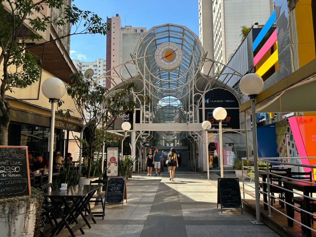 Frente da Rua 24 Horas. Aor redor postes de iluminação e restaurantes. No fundo a entrada com pessoas caminhando. Em cima, uma arquitetura com um relógio.