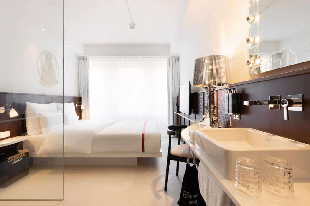 Foto de um quarto do Ruby Claire Hotel Geneva, para ilustrar o post de hotéis em Genebra. Vemos a lateral do quarto. A cama king-size está encostada na parede esquerda. Possui uma mesa de cabeceira com luminária em seus dois lados. No fundo do quarto, está a janela, que ocupa a parede inteira. De frente para a cama, há uma TV fixa na parede. No canto inferior direito da foto, vemos uma bancada branca com uma pia, espelho e abajur.