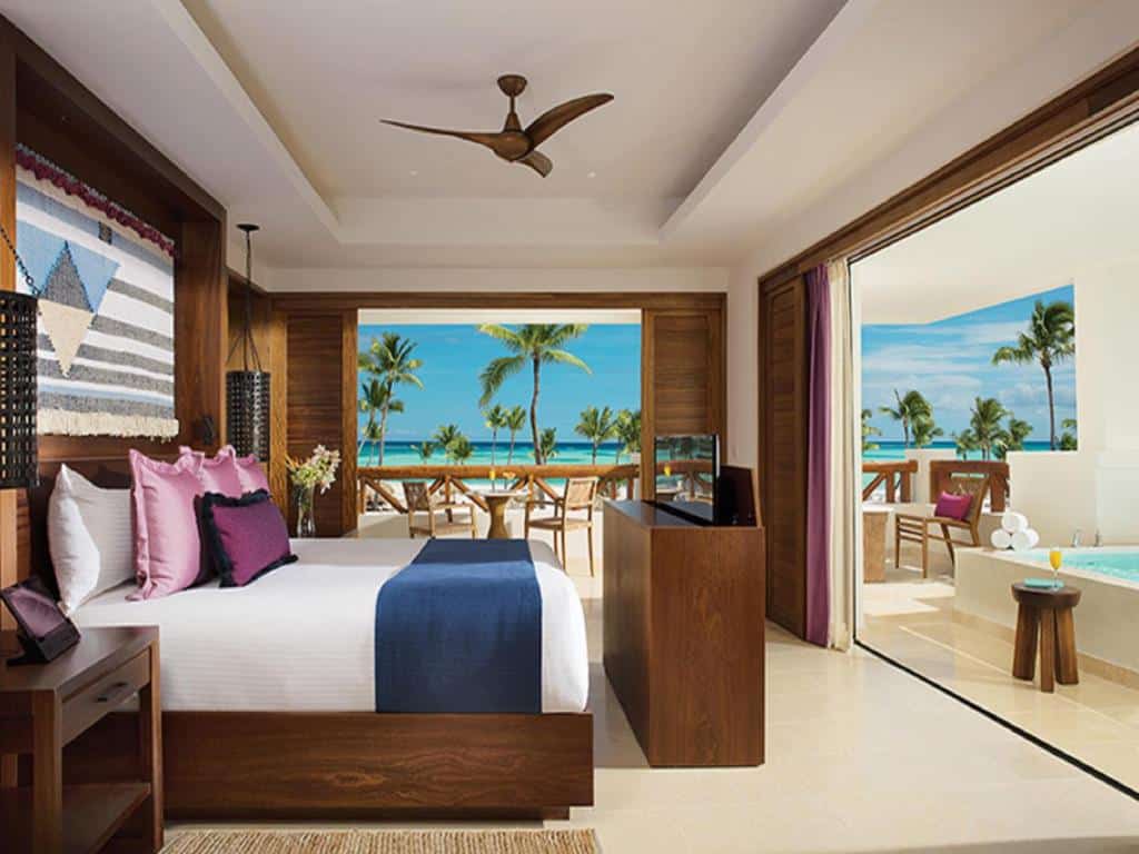 Quarto do Secrets Cap Cana Resort & Spa, um dos melhores hotéis para lua de mel em Punta Cana. A cama de casal está encostada na parede esquerda com mesinhas de cabeceira de madeira dos dois lados e uma cômoda com TV retrátil logo em frente. Há um ventilador de teto acima da cama. Atrás da cômoda e na lateral direita da imagem há uma ampla porta que leva ao terraço, onde há uma banheira de hidromassagem, cadeiras e toalhas. Ao fundo, outra porta leva à varanda com mesa, cadeiras e vista para a praia com palmeiras.