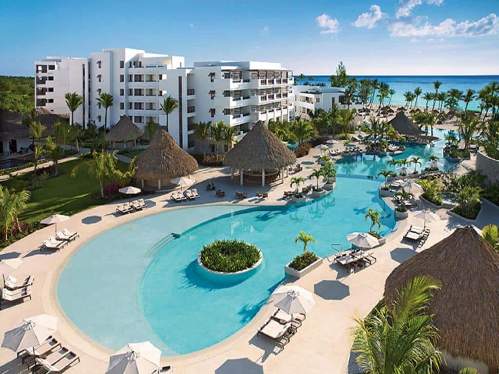 Visão aérea de uma ampla piscina rodeada por espreguiçadeiras e guarda-sóis no Secrets Cap Cana Resort & Spa. Os prédios e bares rodeiam a piscina, e ao fundo é possível ver um pedaçõ do mar e da praia.