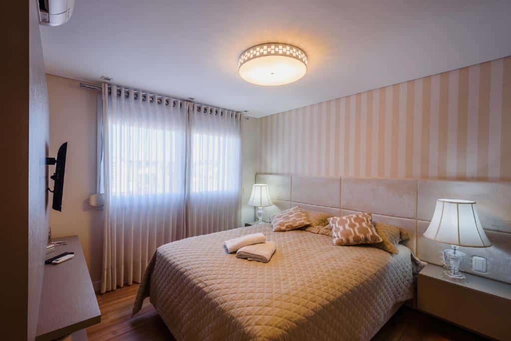 Quarto no Serra Nevada Apartamento. O ambiente é elegante. Há uma cama box de casal no centro, à esquerda. Atrás dela, há uma cabeceira com abajures. Uma janela está do seu lado à esquerda. Na frente da cama há um painel de televisão. Representa o post sobre airbnb em Canela.