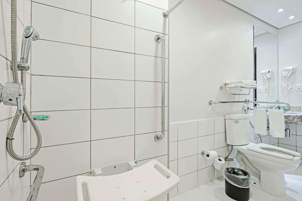 Banheiro com acessibilidade do Slaviero Porto Velho. Do lado direito um vaso sanitário com barras de apoio, o lixo e o papel higiênico. Do lado esquerdo o box com banco e barras de apoio.