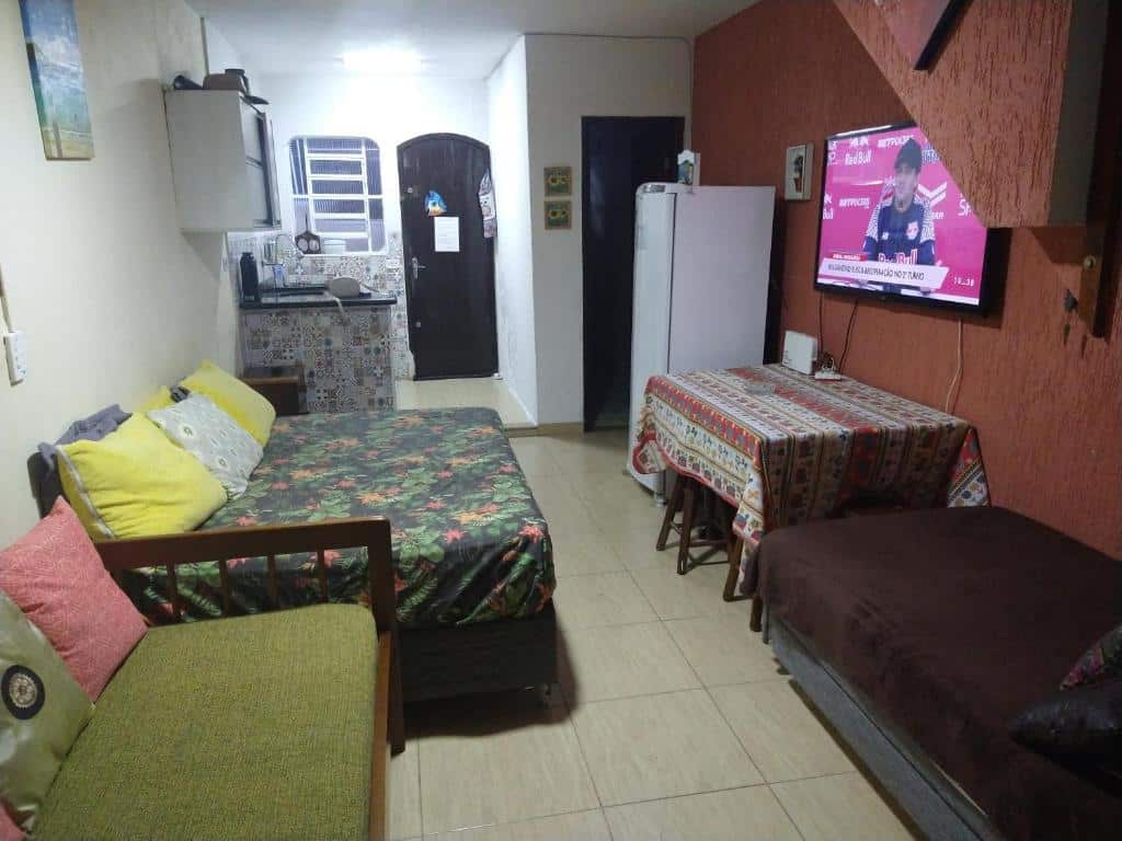 Sala de estar do SOBRADO VILLAGE UBATUBA PER-AÇU. Do lado direito uma cama de solteiro, uma mesa na frente, em cima uma televisão e atrás uma geladeira. Do lado esquerdo um sofá cama, uma cama de casal e no fundo a cozinha.