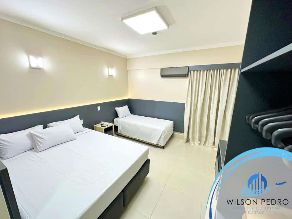 Quarto do Spazzio diRoma. Do lado esquerdo uma cama de casal e uma cama de solteiro, entre elas uma mesinha de apoio com telefone. No fundo uma cortina fechada e um ar-condicionado. Do lado direito um guarda-roupa com cabide.