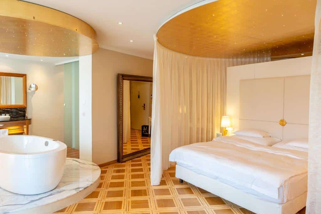 Foto do Hotel des Balances, para representar o post de hotéis em Lucerna. A cama king-size está na direita, e possui cortinas em volta. Do lado esquerdo dessa cortina, há um espelho. Já no canto inferior esquerdo podemos ver de relance a hidro disposta na suíte. O ambiente é espaçoso.
