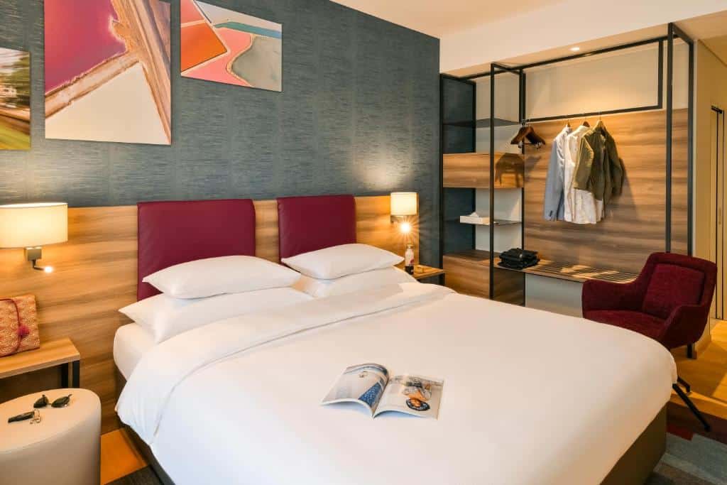Foto de um quarto Mercure Geneva Airport, para ilustrar o post de hotéis em Genebra. O ambiente é moderno. A cama king-size está no centro, possui uma cabeceira atrás com dois abajures e mesa de cabeceira. A parede atrás é em um tom de azul com quadros coloridos. Na direita, há uma estante com araras e cabides para pendurar roupa. Uma poltrona vinho está na frente dessa estante.