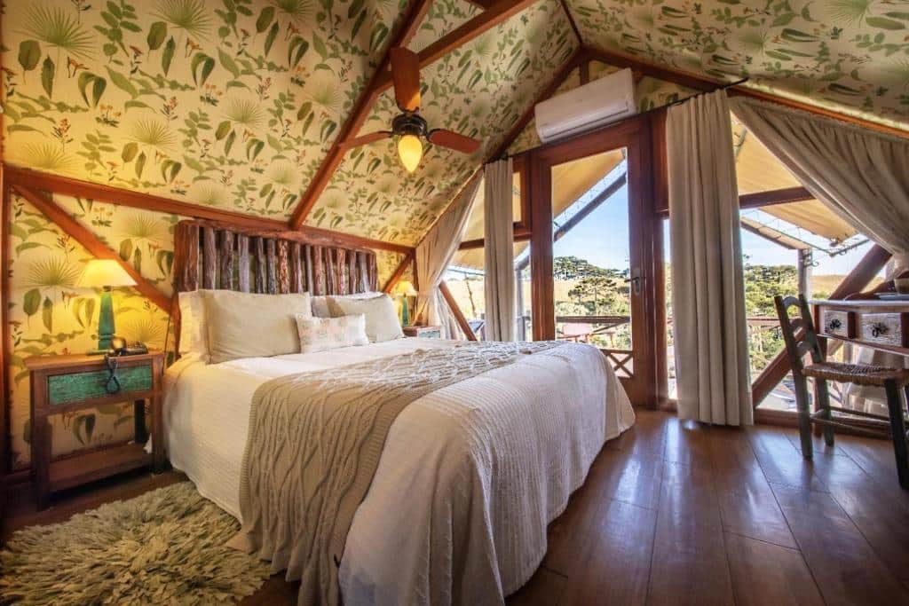 Quarto do hotel Parador Cambará do Sul. A cama está centralizada no quarto e em cada lado da cama há uma pequena mesinha com um abajur em cima. No lado direito da cama há uma parede toda de vidro com uma porta que dá acesso a varanda. Em ferente a cama ainda é possível ver uma mesa com uma cadeira. Imagem para ilustrar o post pousadas em Aparados da Serra.