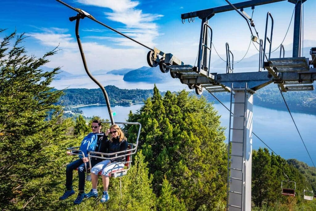Um casal sentado no banco do teleférico, com vista de árvores e um lago no fundo. Imagem para ilustrar post sobre o que fazer em Bariloche.
