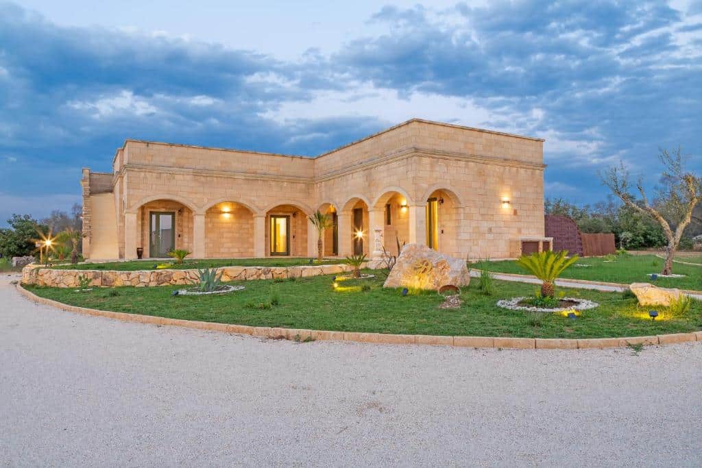 Parte da TENUTA DOROLIVO que mostra um jardim com gramado verde, algumas pedras e plantas e ao fundo uma construção de pedras que forma a hospedagem, ilustrando post Hotéis em Puglia.