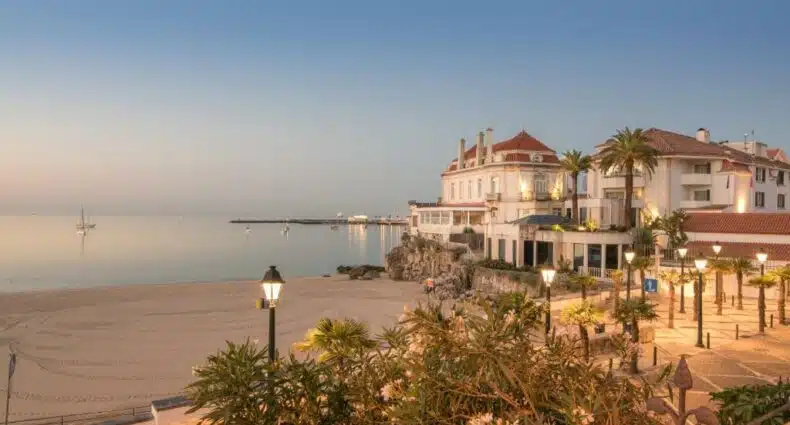 Um calçadão à beira-mar com palmeiras, uma praia de areia e elegantes edifícios do The Albatroz Hotel ao pôr do sol, para representar hotéis românticos perto de Lisboa