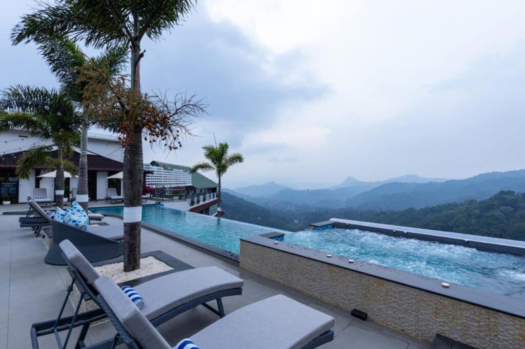 Piscina de borda infinita na cobertura com espreguiçadeiras e palmeiras no The Panoramic Getaway, em Kerala, com vista para uma paisagem montanhosa sob um céu nublado.