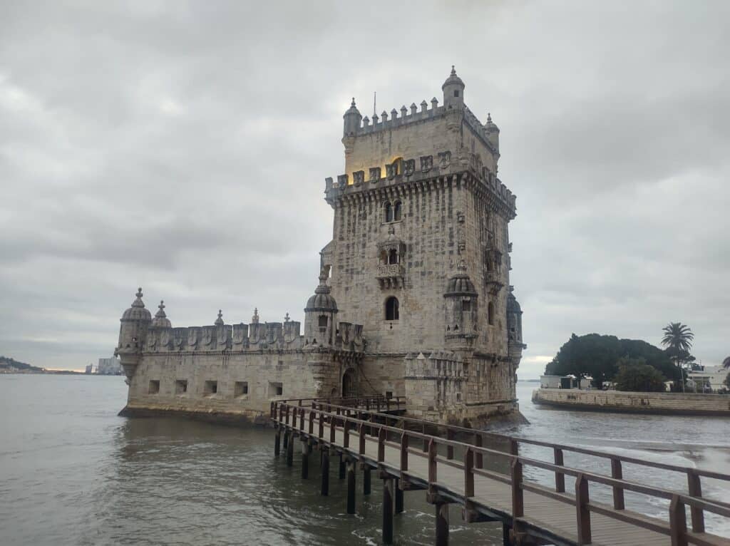Uma fortaleza de pedra com uma torre fica perto da água, conectada à costa por uma passarela de madeira sob um céu nublado, trata-se da Torre de Belém em Lisboa.