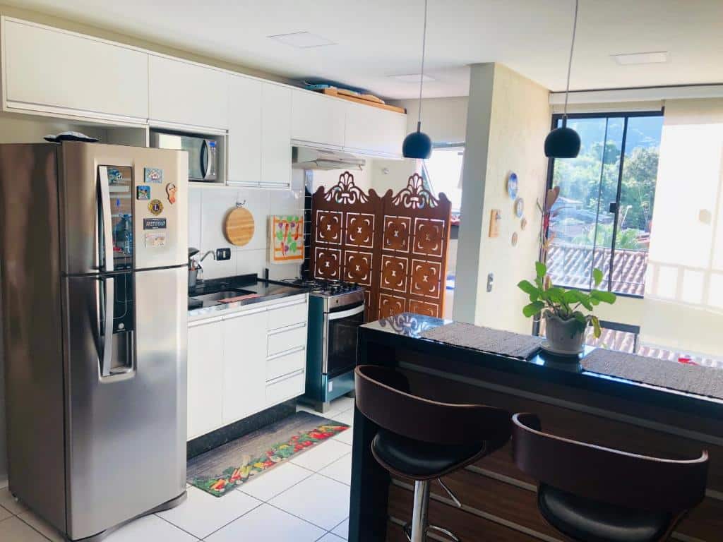 Cozinha do Ubatuba home - Centro. Do lado direito um balcão com cadeira, do lado esquerdo uma geladeira, os armários, o micro-ondas, a pia e o fogão. No fundo as janelas. Foto para ilustrar post sobre airbnb no centro de Ubatuba.