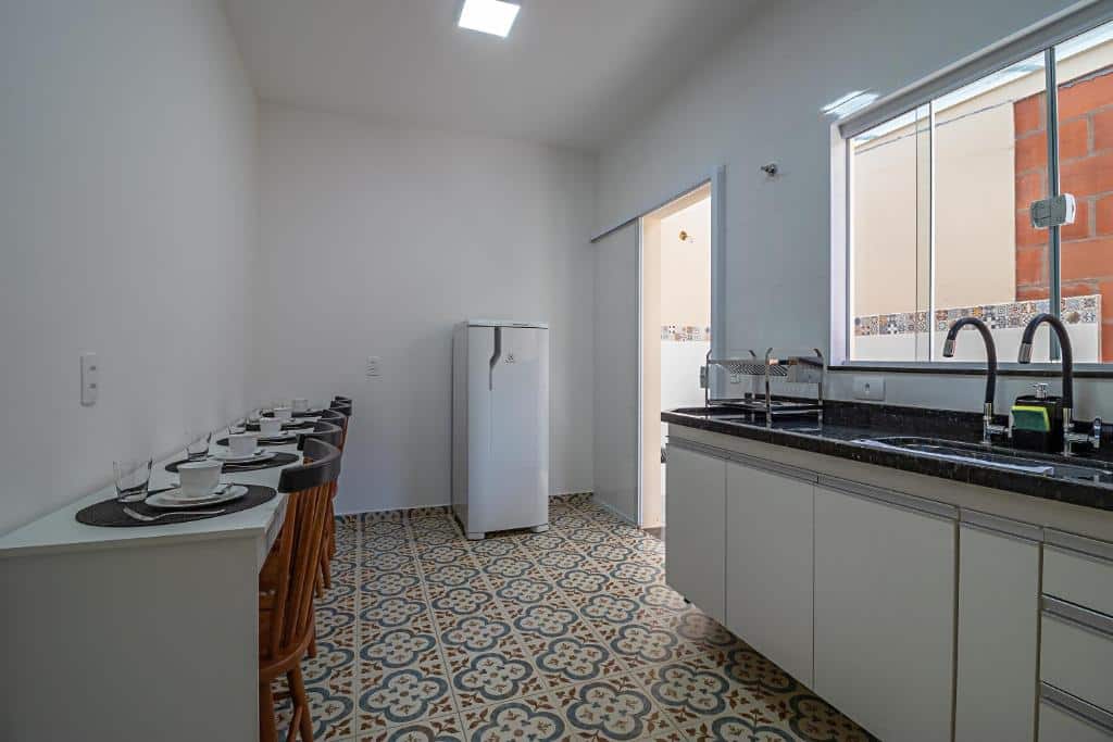 Cozinha do Vila Pepira ViP com uma mesa retangular comprida encostada na parede do lado esquerdo com mesas e utensílios em cima. Já do lado direito tem uma pia para lavar louça com uma janela de vidro. Já ao fundo tem uma geladeira com uma porta de vidro ao lado, imagem ilustrando post Airbnb em Brotas.