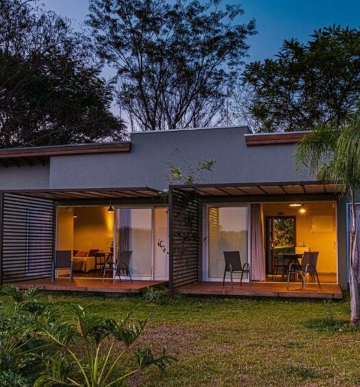 Fachada do Vila Poção como uma casa térrea com duas portas grandes de vidro que mostra a parte interna da casa e cada porta tem uma varanda com cadeiras com vista para as árvores ao redor. Imagem ilustrando post Airbnb em Brotas.