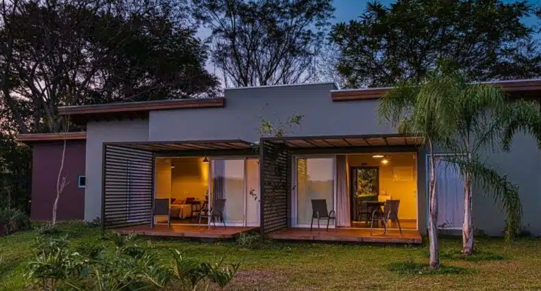 Fachada do Vila Poção como uma casa térrea com duas portas grandes de vidro que mostra a parte interna da casa e cada porta tem uma varanda com cadeiras com vista para as árvores ao redor. Imagem ilustrando post Airbnb em Brotas.