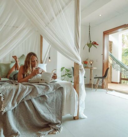 Mulher loira deitada em cima de uma cama enquanto lê um livro. Está em um quarto no bungalow, que possui uma varanda com rede à direita.
