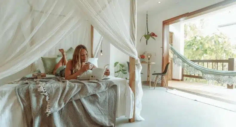 Mulher loira deitada em cima de uma cama enquanto lê um livro. Está em um quarto no Villa Estrela Bungalow, que possui uma varanda com rede à direita, para representar aibnb em Itacaré.
