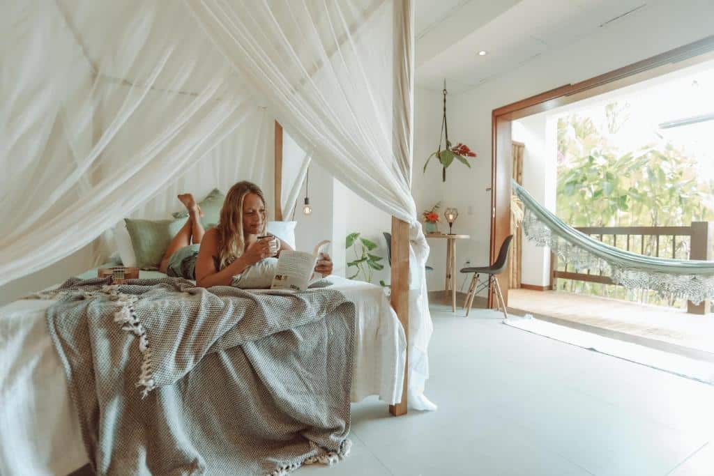 Mulher loira deitada em cima de uma cama enquanto lê um livro. Está em um quarto no bungalow, que possui uma varanda com rede à direita.