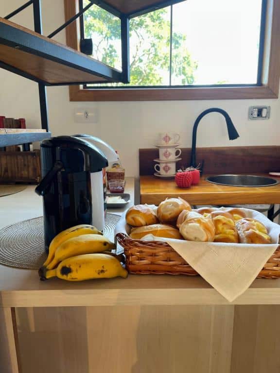 Balcão do Villa Itália. Um balcão com uma cesta com pães, bananas e duas garrafas térmicas. No fundo, uma pia e xícaras.