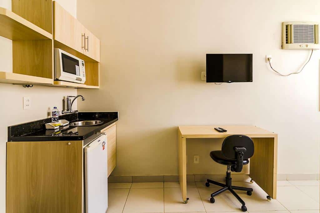 Outro ângulo no quarto da Villa Neves Residence.  Na direita há uma mesa com cadeira giratória e TV acima, fixa na parede. Na esquerda, uma mini cozinha.