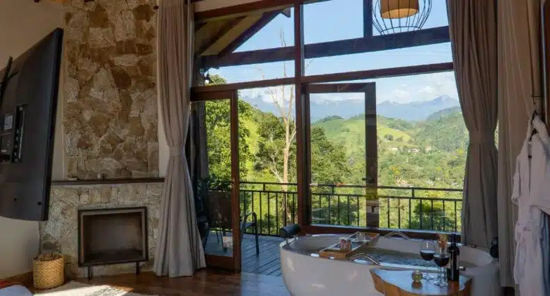 Imagem do quarto em Monte Mauá Chalés, que está ilustrando o post sobre airbnb em Visconde de Mauá. Há uma cama box de casal no centro do quarto, com pétalas de rosa espalhada por cima dela e do chão do quarto. Do lado direito da cama há uma lareira, varanda e também uma banheira. O ambiente é espaçoso. Uma TV está fixa em uma coluna de frente para a cama.