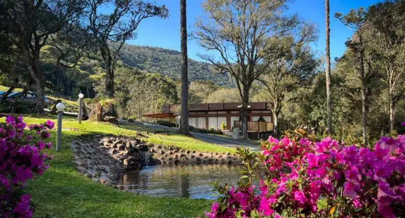A imagem mostra uma paisagem tranquila e natural em frente à Pousada Encanto do Avencal, um dos chalés na Serra Catarinense. No primeiro plano, flores vibrantes cor-de-rosa (provavelmente azaleias) adornam o lado direito da foto, emoldurando a cena com um toque de cor. No centro, um pequeno lago cercado por pedras, com uma suave queda d'água, adiciona uma sensação de serenidade. Ao fundo, destacam-se árvores altas e montanhas verdes, sob um céu azul claro. A pousada, com sua arquitetura rústica e elegante, possui linhas simples, mas refinadas, misturando-se perfeitamente ao ambiente natural. O design acolhedor e sofisticado, junto com a beleza da paisagem ao redor, cria uma atmosfera de charme e conforto.