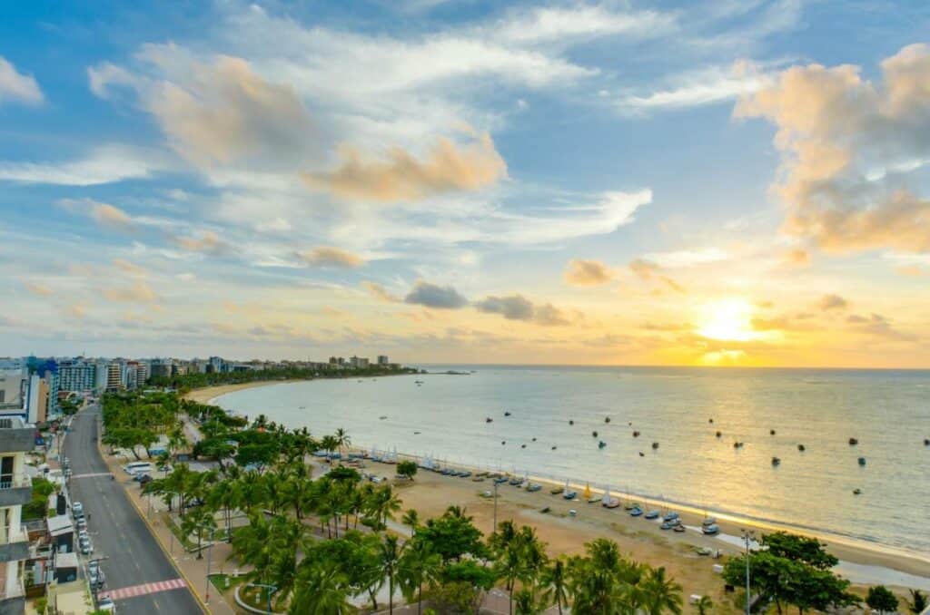 Vista da praia Jatiúca pelo Meridiano Hotel, onde ficar em Maceió. Há um pôr do sol refletindo no mar, que tem vários barcos navegando. Há várias árvores na orla da paia, que tem uma avenida com carros estacionados