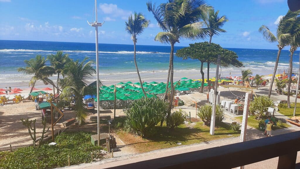 Vista da praia do Francês de um quarto do hotel Ponta Verde Francês. A praia tem vários guarda-sóis abertos com mesas e cadeiras espalhadas na areia, algumas árvores e o mar com poucas ondas