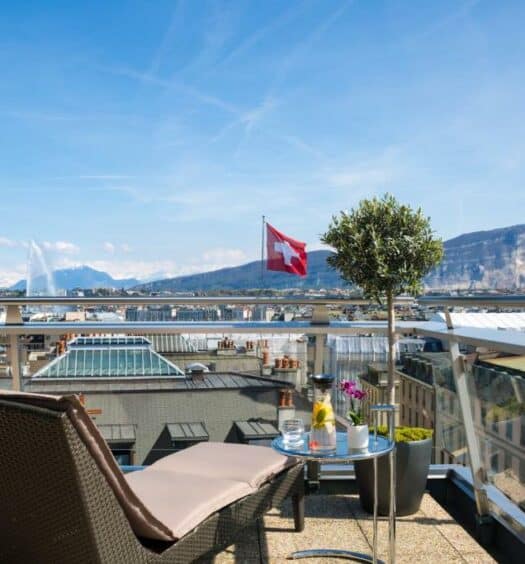 Terraço do Warwick Geneva. O céu está limpo e azul. Há uma espreguiçadeira e uma mesa redonda circular no seu lado direito, para representar hotéis em Genebra.