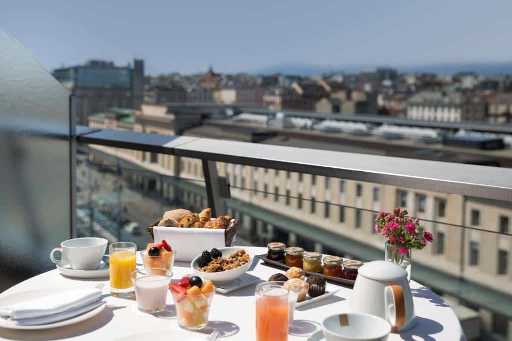 Foto tirada num terraço do Warwick Geneva, de uma mesa com café da manhã. As opções são de cereais, croissants, geleias, sucos, frutas, etc. A vista é para a cidade de Genebra.