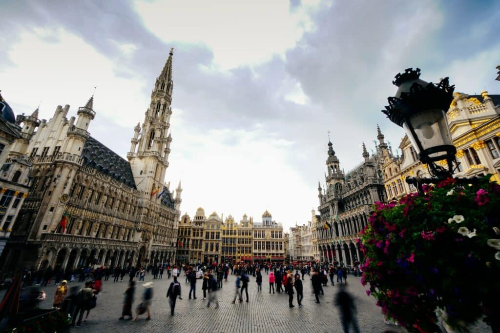 Praça do Luxemburgo em Bruxelas, trata-se de um local com diversos prédios medievais e históricos, muitos com torres, outros com detalhes em dourado, no pátio que fica no centro do local há muitas pessoas caminhando