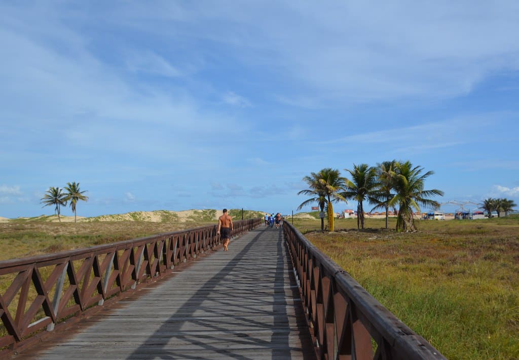 Calçadão de madeira com pessoas caminhando, palmeiras à direita, área gramada e céu azul com nuvens, na Praia de Atalaia em Aracaju.