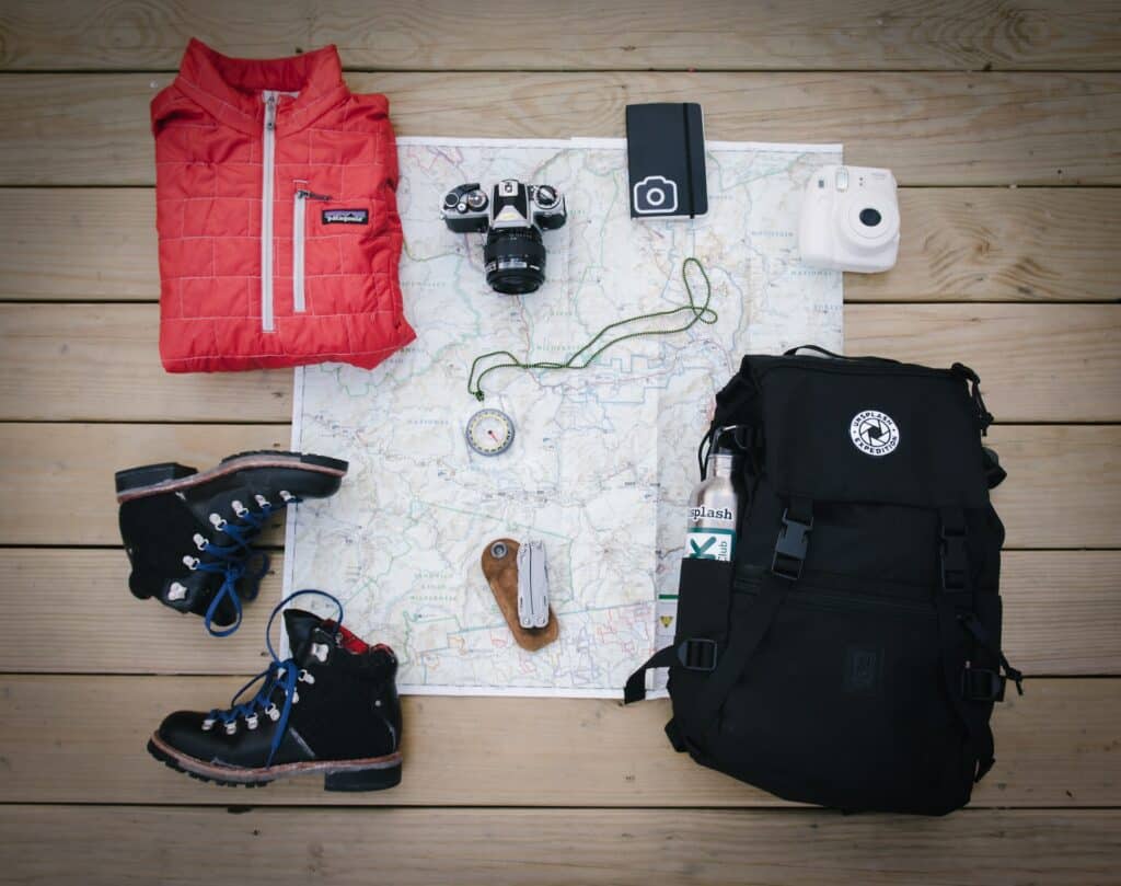 Uma mochila preta, um mapa, uma bússola, um par de botas, duas câmeras fotográficas e um casaco, todos os itens apoiados em um chão de madeira, para representar seguro viagem para quem viaja muito
