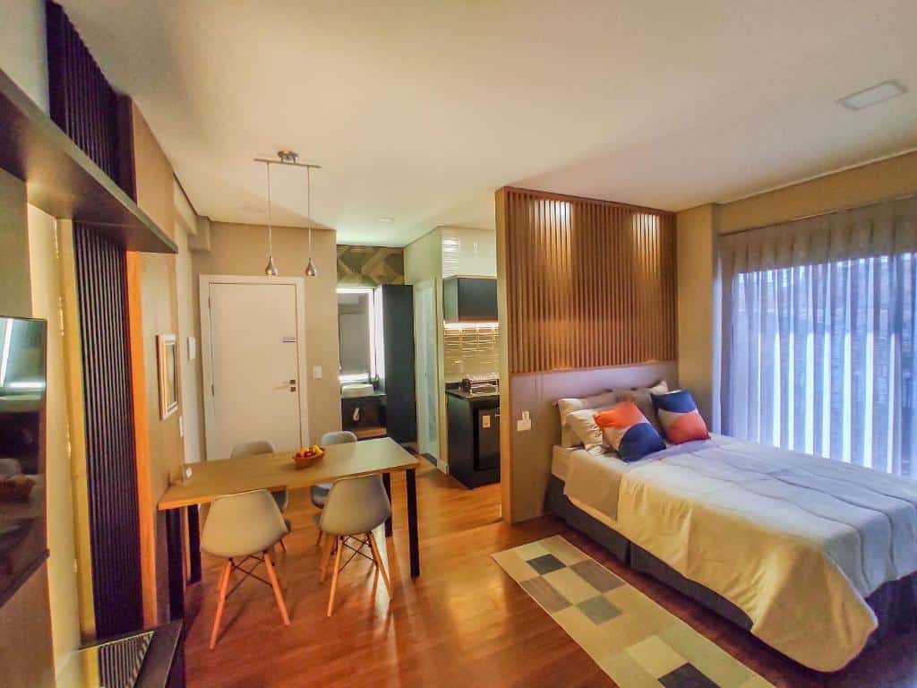 Foto do Apartamentos Studio no centro de Foz do Iguaçu. A cama está na direita, e do lado direito da cama há uma janela, e atrás, um painel que a separa da cozinha. Do lado esquerdo da cama há uma mesa com quatro cadeiras.