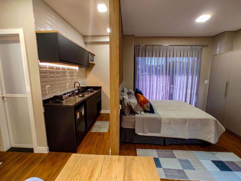 Outro ângulo do Apartamentos Studio no centro de Foz do Iguaçu, onde vemos de lado a cama e a cozinha atrás dela, que tem armários, pia, frigobar, cooktop e micro-ondas.