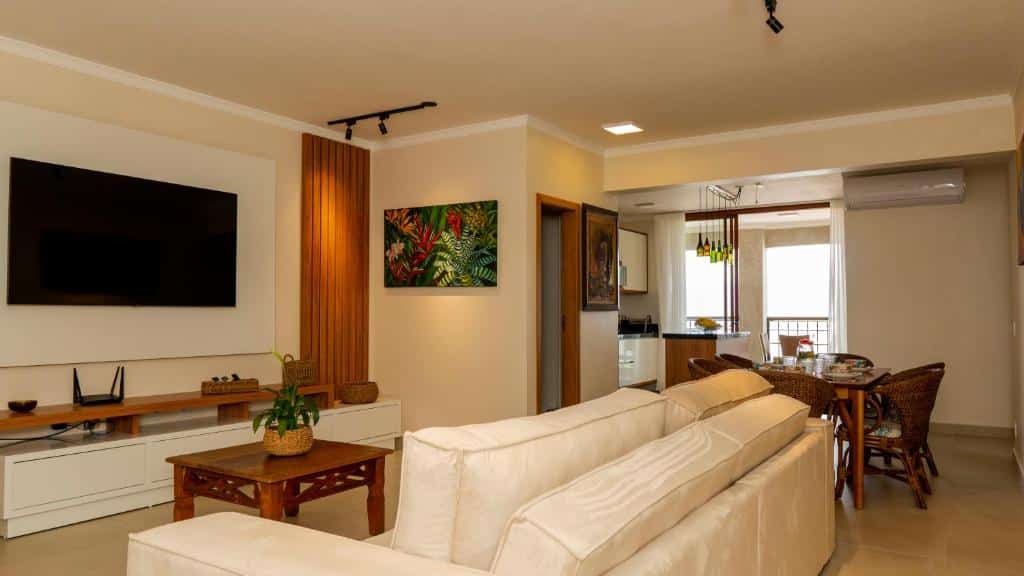 Sala de estar do Apartamento de Frente para o Mar no Itagua. Um sofá na frente, no lado esquerdo uma mesa de centro, a raque e a televisão. No fundo, no lado direito uma mesa, em cima ar-condicionado. Atrás a cozinha.