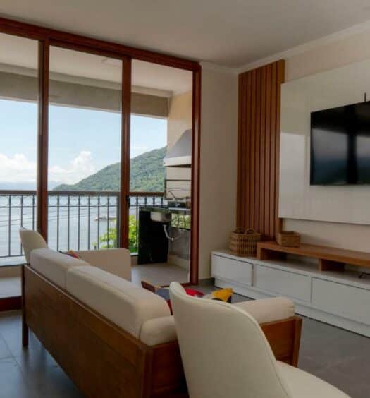 Foto da sala de estar do APARTAMENTO DE FRENTE PARA O MAR para ilustrar post sobre airbnb em Itaguá. No fundo uma varanda com vista para o mar, no lado direito um painel com televisão, e no meio um sofá com poltrona.