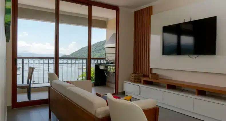 Foto da sala de estar do APARTAMENTO DE FRENTE PARA O MAR para ilustrar post sobre airbnb em Itaguá. No fundo uma varanda com vista para o mar, no lado direito um painel com televisão, e no meio um sofá com poltrona.