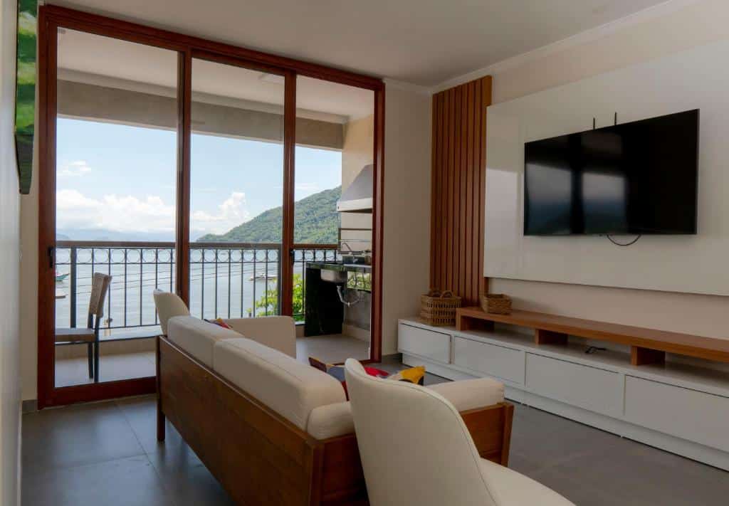 Foto da sala de estar do APARTAMENTO DE FRENTE PARA O MAR para ilustrar post sobre airbnb em Itaguá. No fundo uma varanda com vista para o mar, no lado direito um painel com televisão, e no meio um sofá com poltrona.