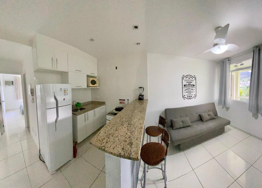 Sala e cozinha do airbnb Eco Ribeira. No lado direito da imagem há uma janela com cortinas, um sofá e um balcão que divide o cômodo com duas banquetas. No lado direito do balcão há um fogão, uma bancada com pia e armário, em cima da pia outro armário com um microondas e no lado esquerdo da pia uma geladeira. Ainda no lado esquerdo do cômodo há um corredor que dá acesso ao quarto.