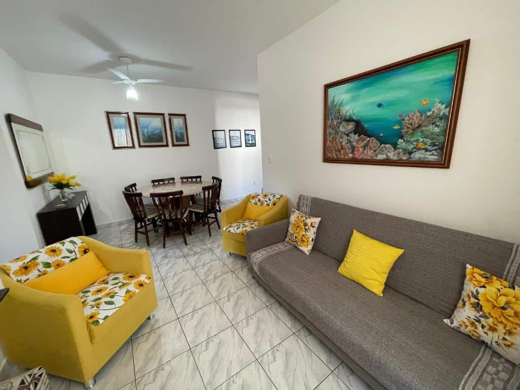 Sala de estar do Apartamento Ubatuba, Itaguá. Do lado esquerdo uma poltrona, no fundo uma estante e um espelho em cima. No meio uma mesa com cadeiras. Do lado direito um sofá e uma poltrona.