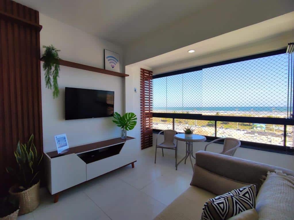 Sala de estar do Apartamento vista mar Atalaia. Um sofá do lado direito, de frente uma raque e uma televisão. No fundo uma mesa com duas cadeiras e a janela da sacada com vista para o mar.