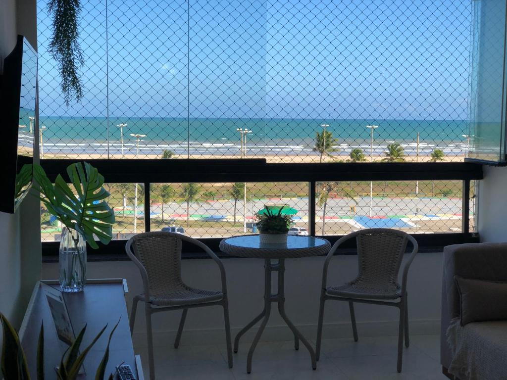 Sala de estar do Apartamento vista mar Atalaia. Do lado direito um sofá, do lado esquerdo a raque com TV. No meio uma mesa com duas cadeiras e uma parede de vidro com vista para a praia.