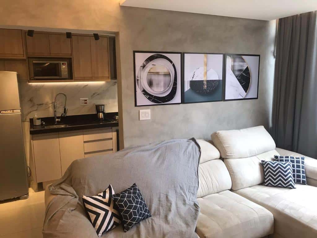 Apto aconchegante vista mar. Um sofá com manta e travesseiros na frente, atrás uma cozinha completa.