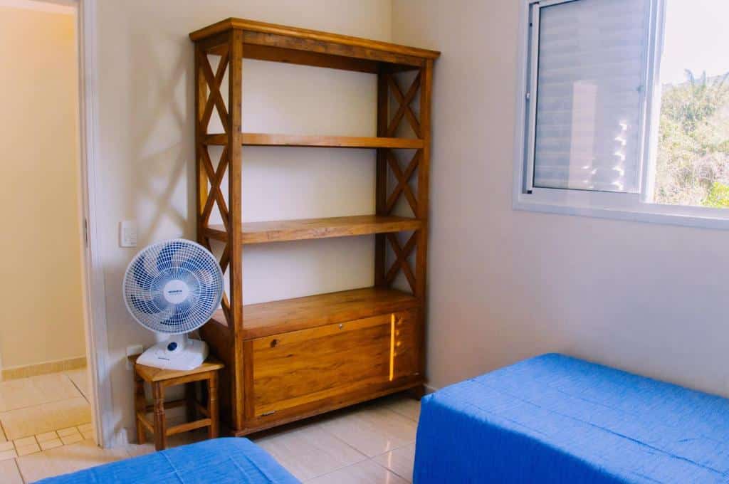Quarto do Apto no Itaguá. Duas camas de solteiro de frente para um ventilador em cima de um banco e uma estante de madeira. Do lado esquerdo a porta do quarto, do lado direito a janela. Foto para ilustrar post sobre airbnb em Itaguá.