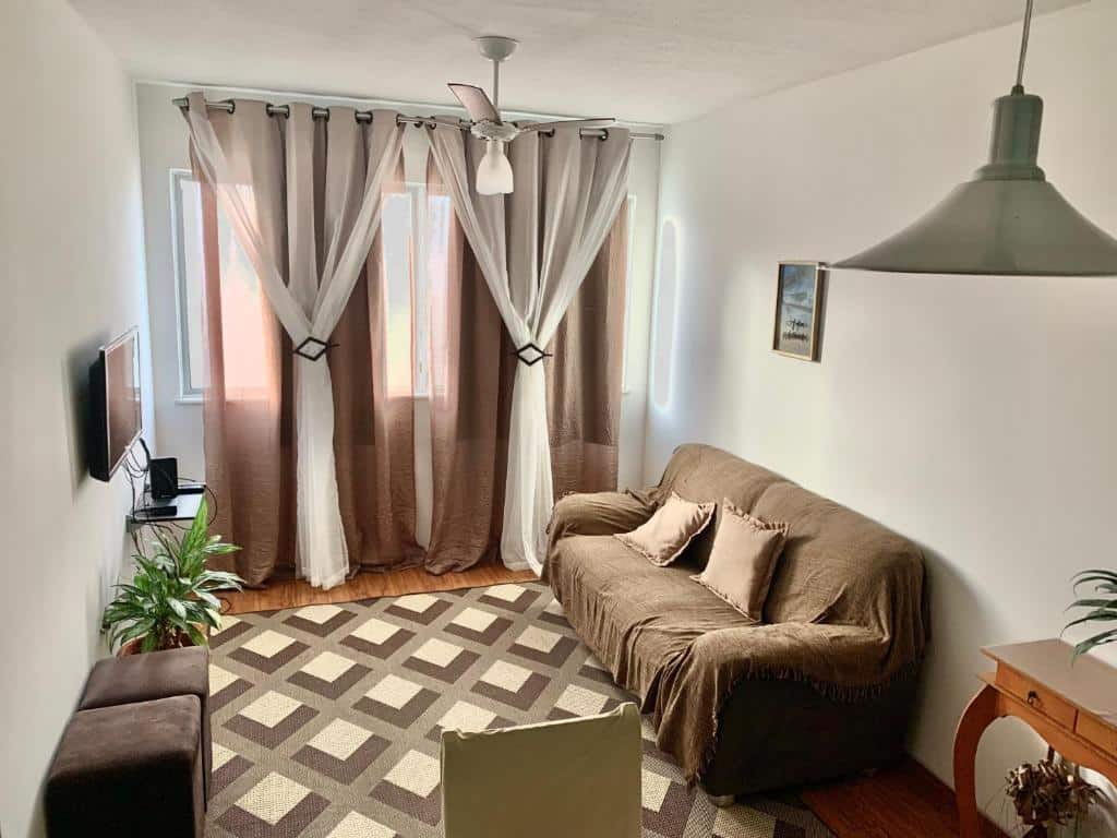 Sala de estar do Apto Praia Atalaia. Na frente uma mesa com cadeiras e um abajur em cima. Do lado direito um sofá, de frente uma televisão, uma prateleira, puff e um tapete. No fundo uma janela com cortinas.