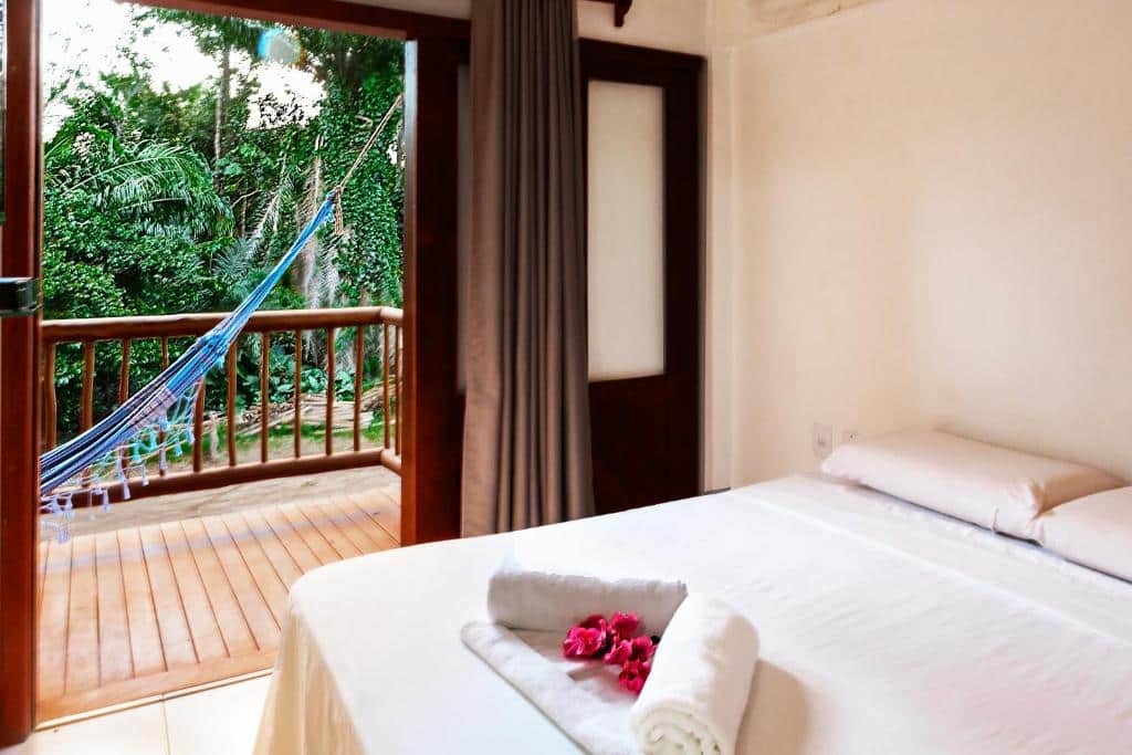 Quarto no Arpoador Flat. Há uma cama de casal no canto direito na foto, enquanto no esquerdo está a varanda com vista e uma rede de descanso. Representa o post sobre airbnb em Itacaré.
