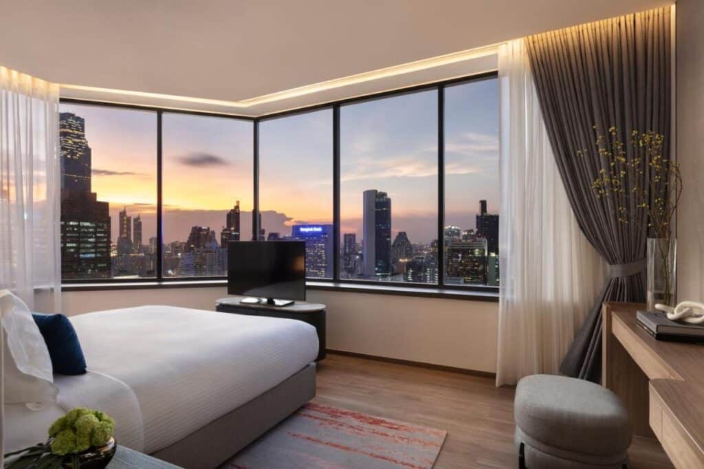 Quarto do hotel Ascott Embassy Sathorn com cama grande, TV e escrivaninha. Janelas do chão ao teto revelam o horizonte da cidade ao pôr do sol.
