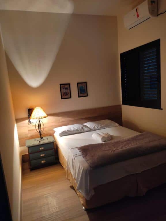 Quarto no AVENIDA HOTEL DE RESENDE. Há uma cama de casal e uma mesa de escrivaninha com abajur na sua esquerda. À direita, há uma janela. É uma indicação de hotéis perto da Dutra.